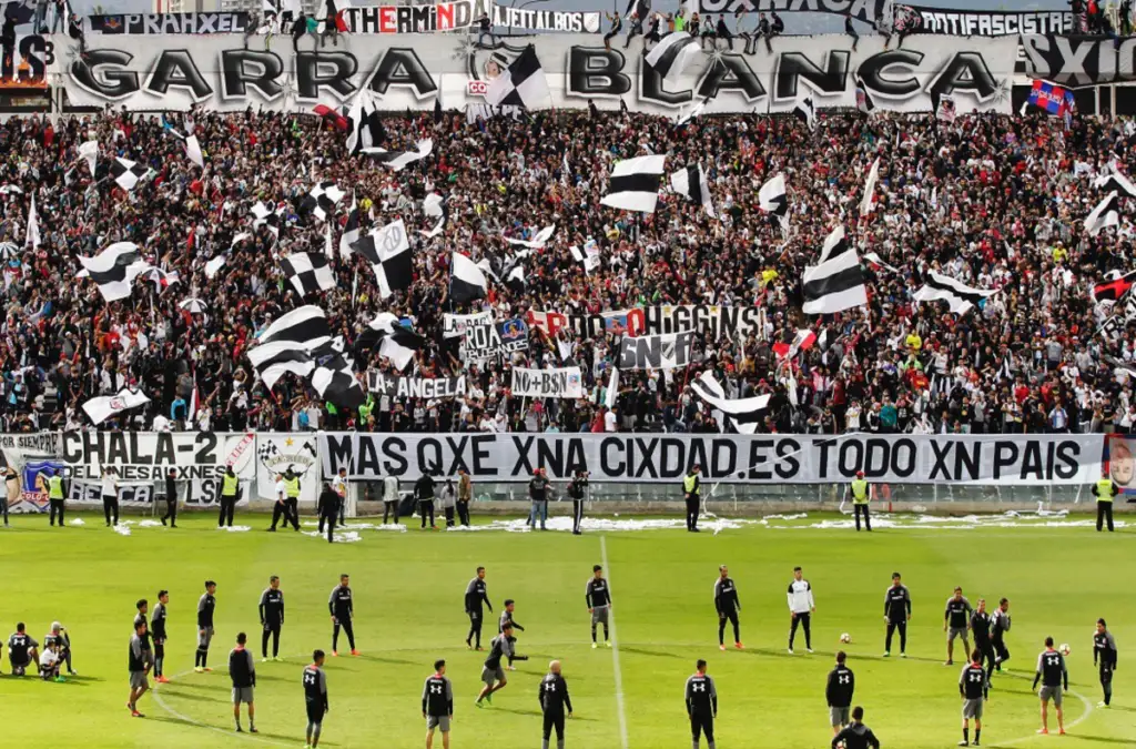 Hinchas de Colo-Colo desplegados en el sector cordillera del Estadio Monumental, observando un entrenamiento de Colo-Colo en la cancha principal del recinto deportivo de Macul. En la parte posterior de la galería se puede observar un lienzo con la consigna «Garra Blanca».