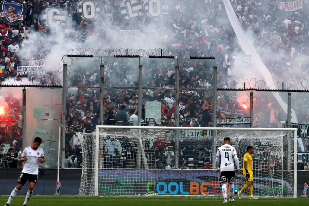 Fotografía a la galería norte del Estadio Monumental, donde se puede ver repleta de hinchas que encienden bengalas y levantan los bombos para alentar a los jugadores de Colo-Colo en pleno partido. En el césped se puede ver en miniatura a Alan Saldivia, Brayan Cortés y Esteban Pavez.