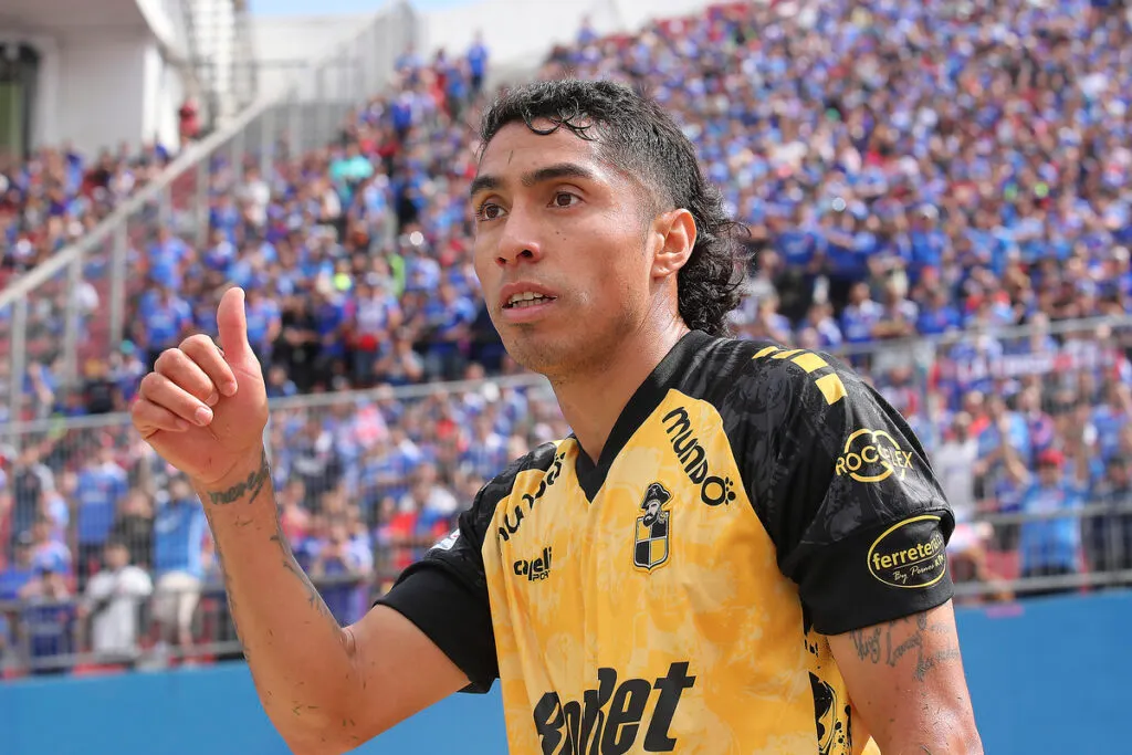 Luciano Cabral levanta su pulgar en pleno partido con l camiseta de Coquimbo Unido y la hinchada de Universidad de Chile de fondo.